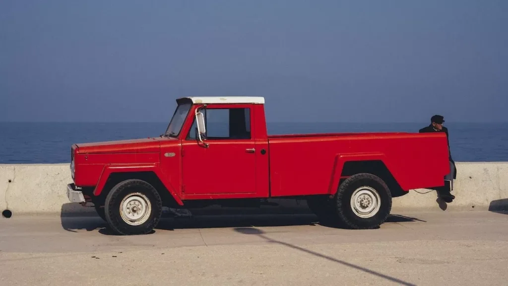 Classic red pickup truck parked near the ocean with a person sitting nearby