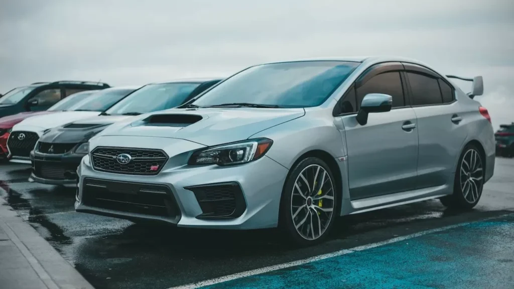A silver Subaru WRX parked in a city parking lot with other cars on a rainy day