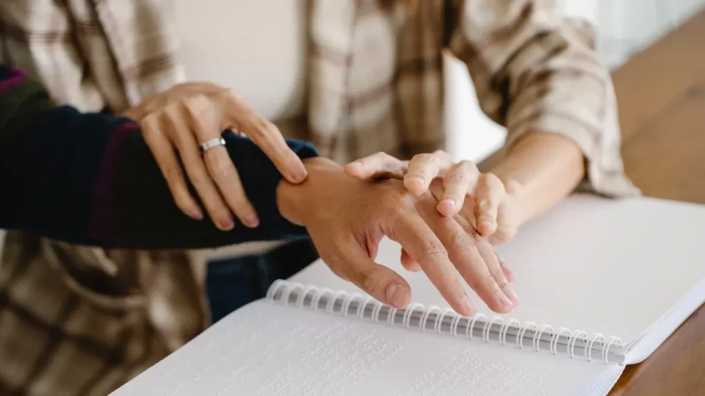Two people reading a Braille book, focusing on hands guiding and assisting in touch-reading.