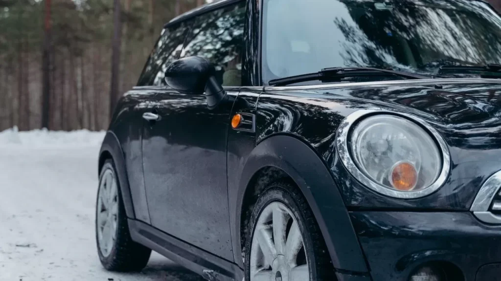 A sleek black car parked on a snowy forest road during winter, surrounded by tall pine trees