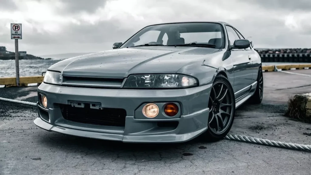 A classic gray sports car parked on a waterfront under a gloomy sky