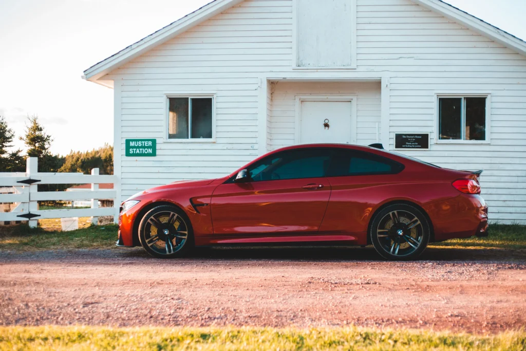A sleek red luxury coupe car parked outside a rustic white house on a sunny day