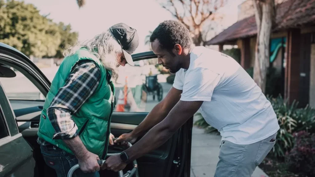 Young volunteer helping senior man with walker near a parked car