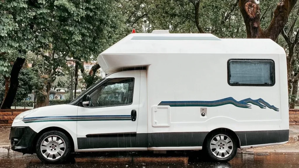 Camper van parked on a rainy street surrounded by lush green trees, reflecting on wet asphalt