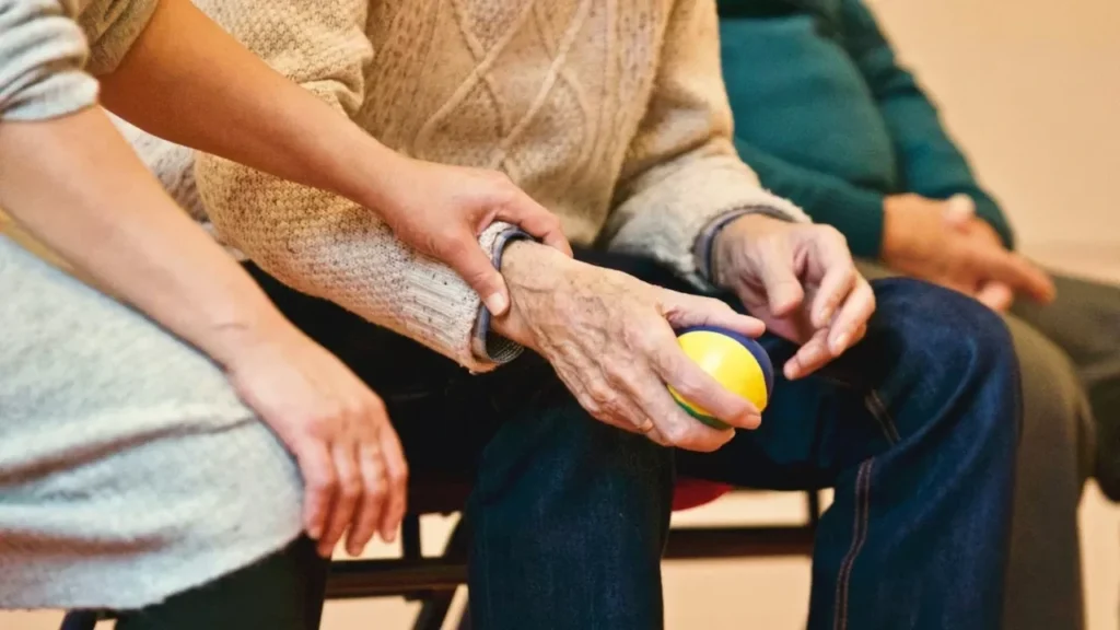 An elderly person receives support from a caregiver, holding hands indoors