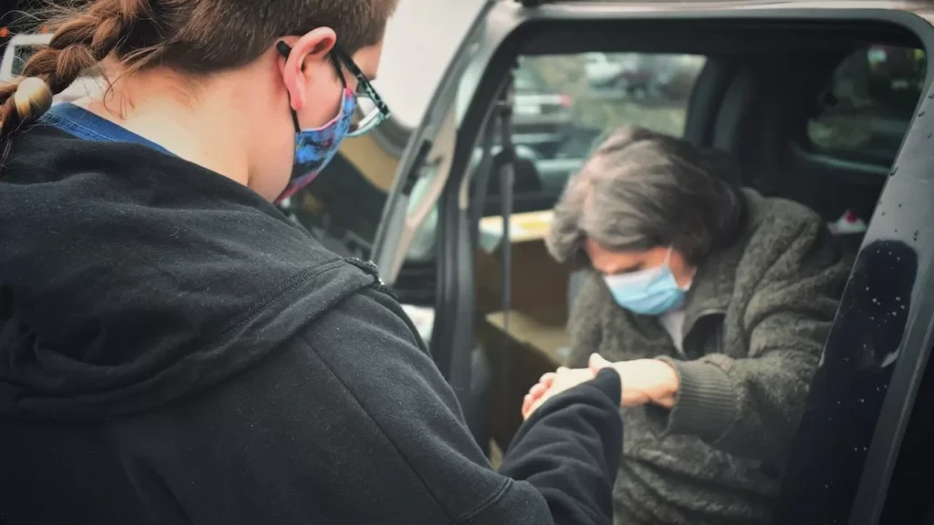 A person assists an elderly man wearing a mask by their car, symbolizing care during the pandemic