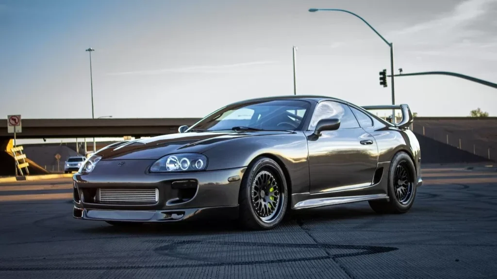 A sleek Toyota Supra sports car is parked on an outdoor asphalt roadway with a sunset backdrop