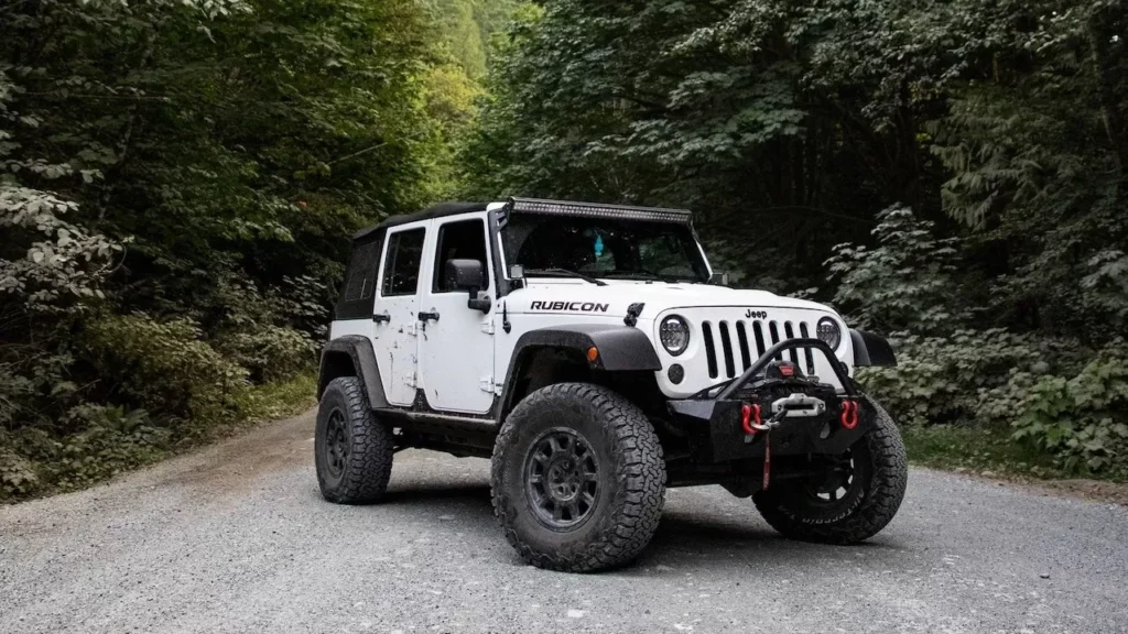 A rugged Jeep Wrangler on a gravel road surrounded by lush forest and towering mountains