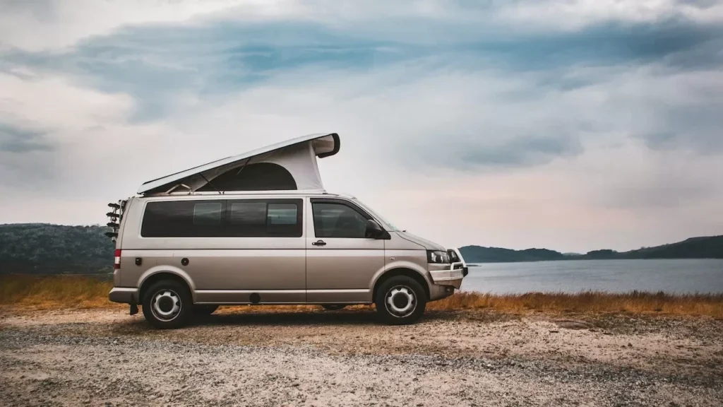 A camper van with rooftop tent parked by a tranquil lake under cloudy skies