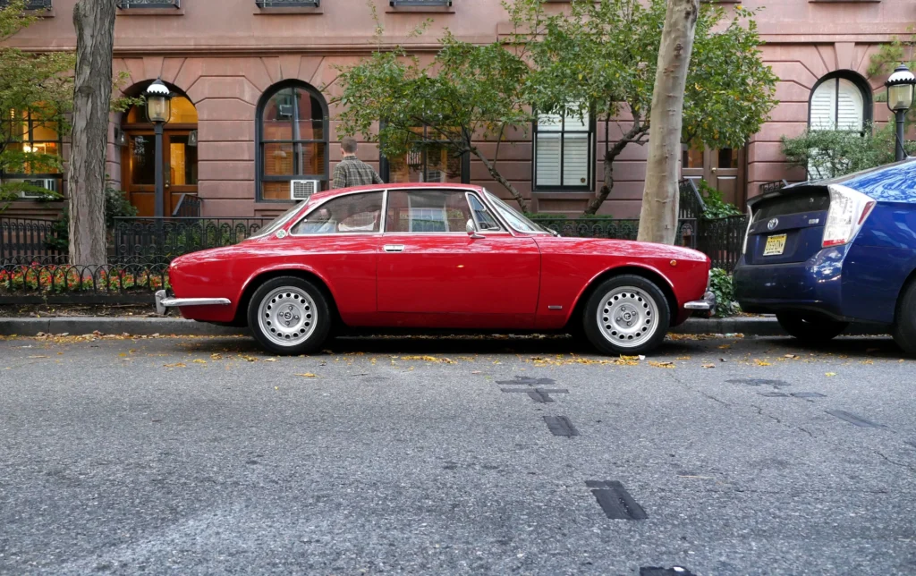 A classic red car parked on a city street with trees and buildings in the background