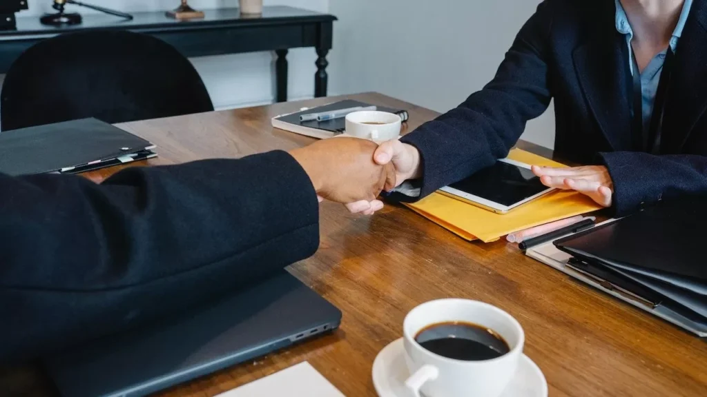 Professional business meeting handshake over a table with coffee, documents, and tech devices in an office setting