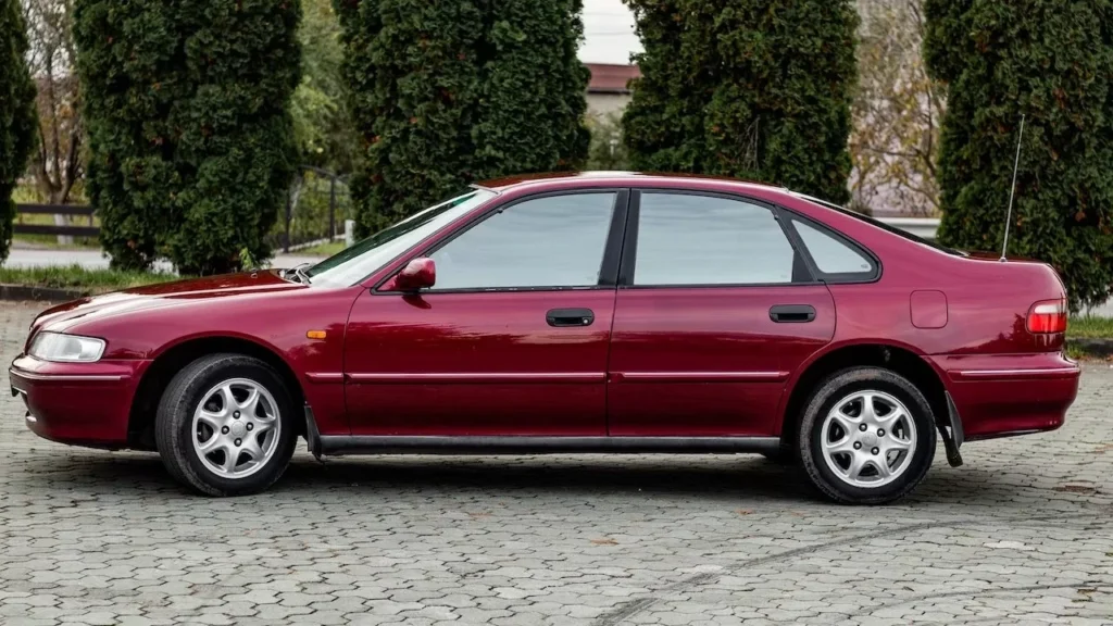 Side view of a red sedan parked on a paved area, surrounded by tall trees