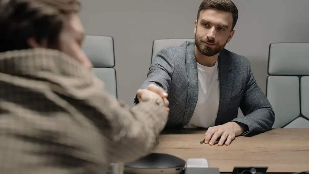 Two businessmen shaking hands across a wooden desk in a modern office setting