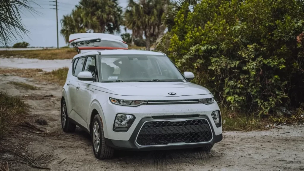 A white Kia Soul X-Line parked on a dirt road surrounded by lush greenery, featuring a roof cargo box